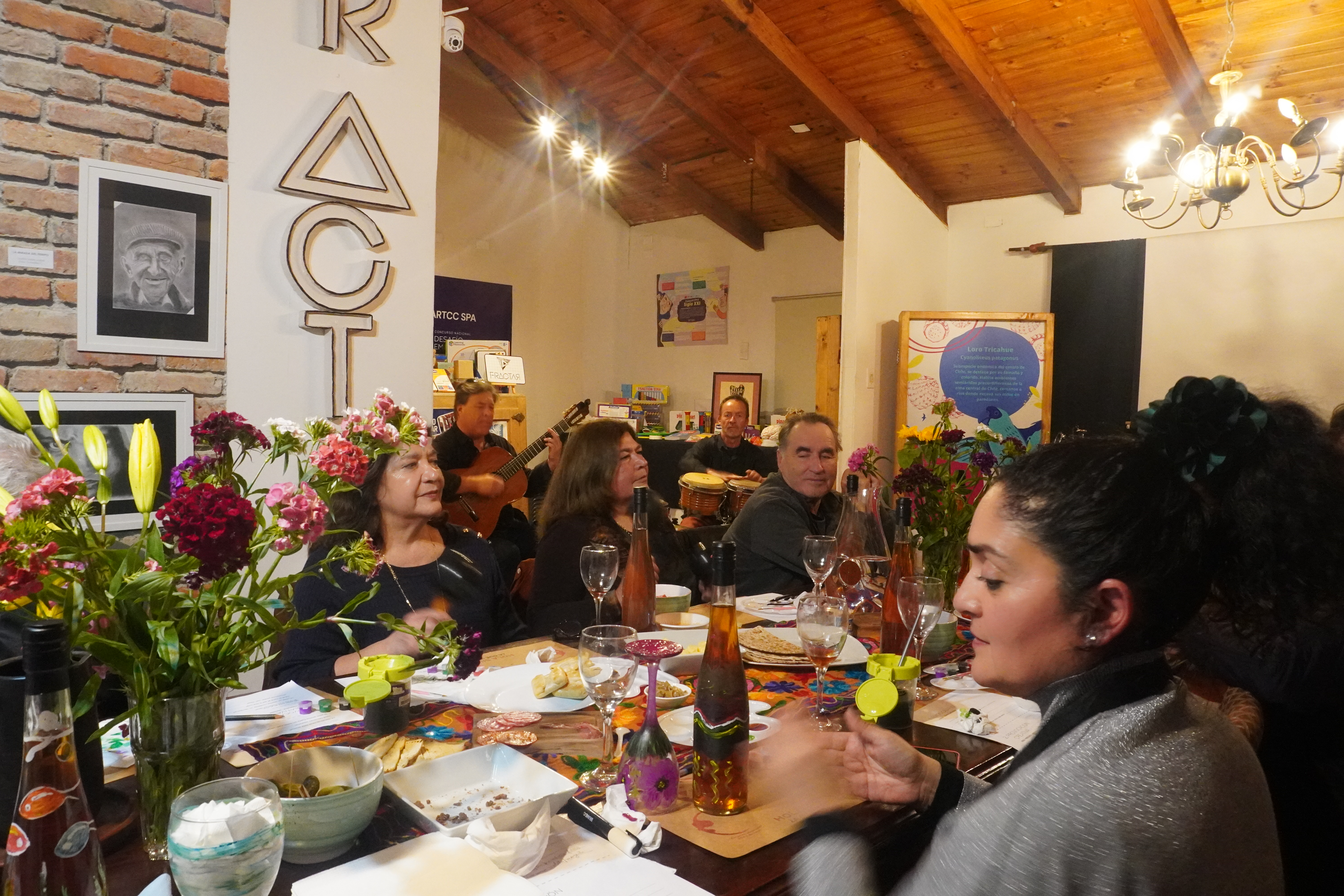 Personas reunidas alrededor de una mesa con músicos tocando guitarra y percusión al fondo